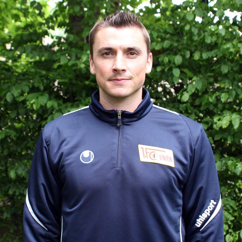 Mann in sportlicher Jacke mit Logo steht vor einem Hintergrund aus grünen Blättern.