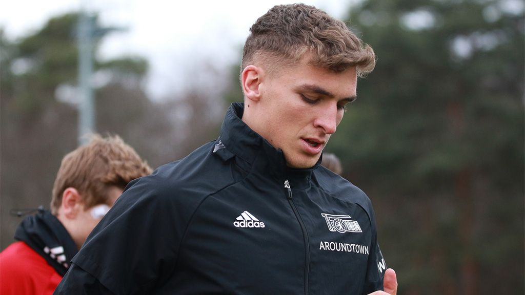 A young man in a black sports jacket, looking thoughtful and focused on something. Blurry figures are in the background.