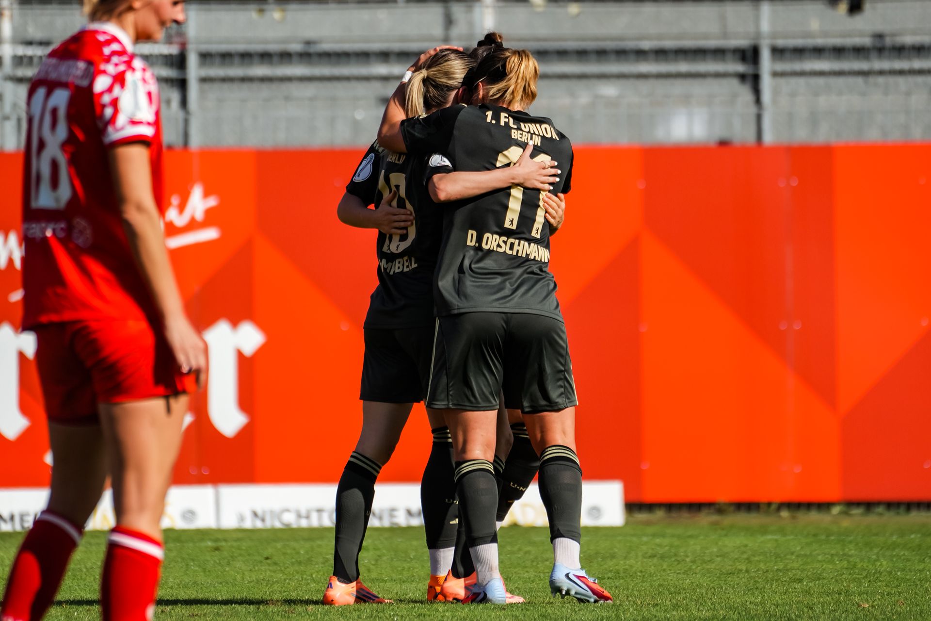 Drei Fußballspielerinnen in schwarzen Trikots umarmen sich auf dem Spielfeld nach einem Erfolg. Im Hintergrund ist eine Werbetafel sichtbar.
