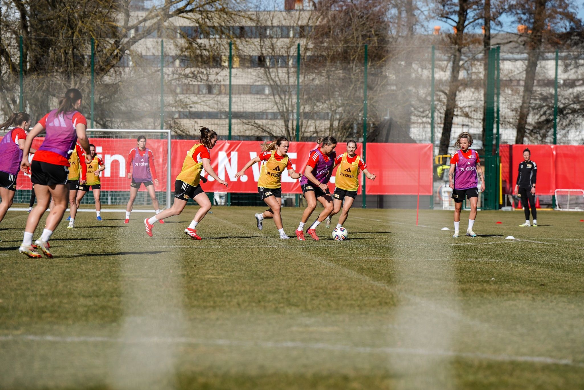 Frauenfußballmannschaft trainiert auf dem Platz. Spielerinnen dribbeln und passen den Ball. Sportliche Kleidung und ein Stadion im Hintergrund.