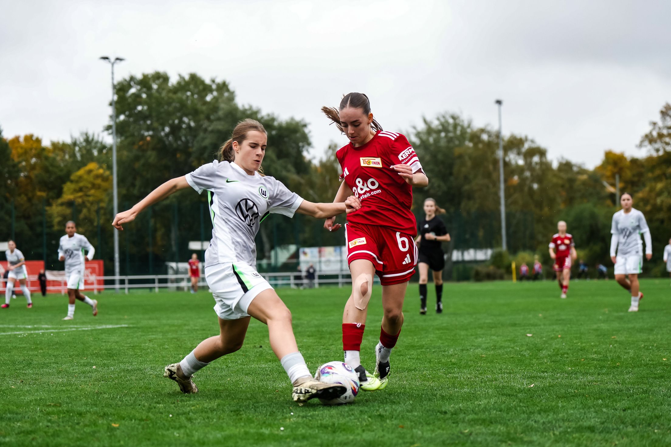 Zwei Frauen im Fußballtrikot kämpfen um den Ball auf einem Rasenplatz während eines Spiels. Wolkiger Himmel im Hintergrund.