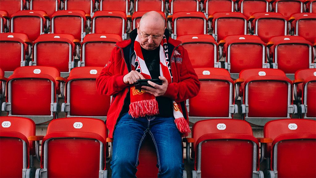 Ein Mann in einem roten Schal sitzt allein zwischen leeren roten Stadionplätzen und schaut auf sein Handy.