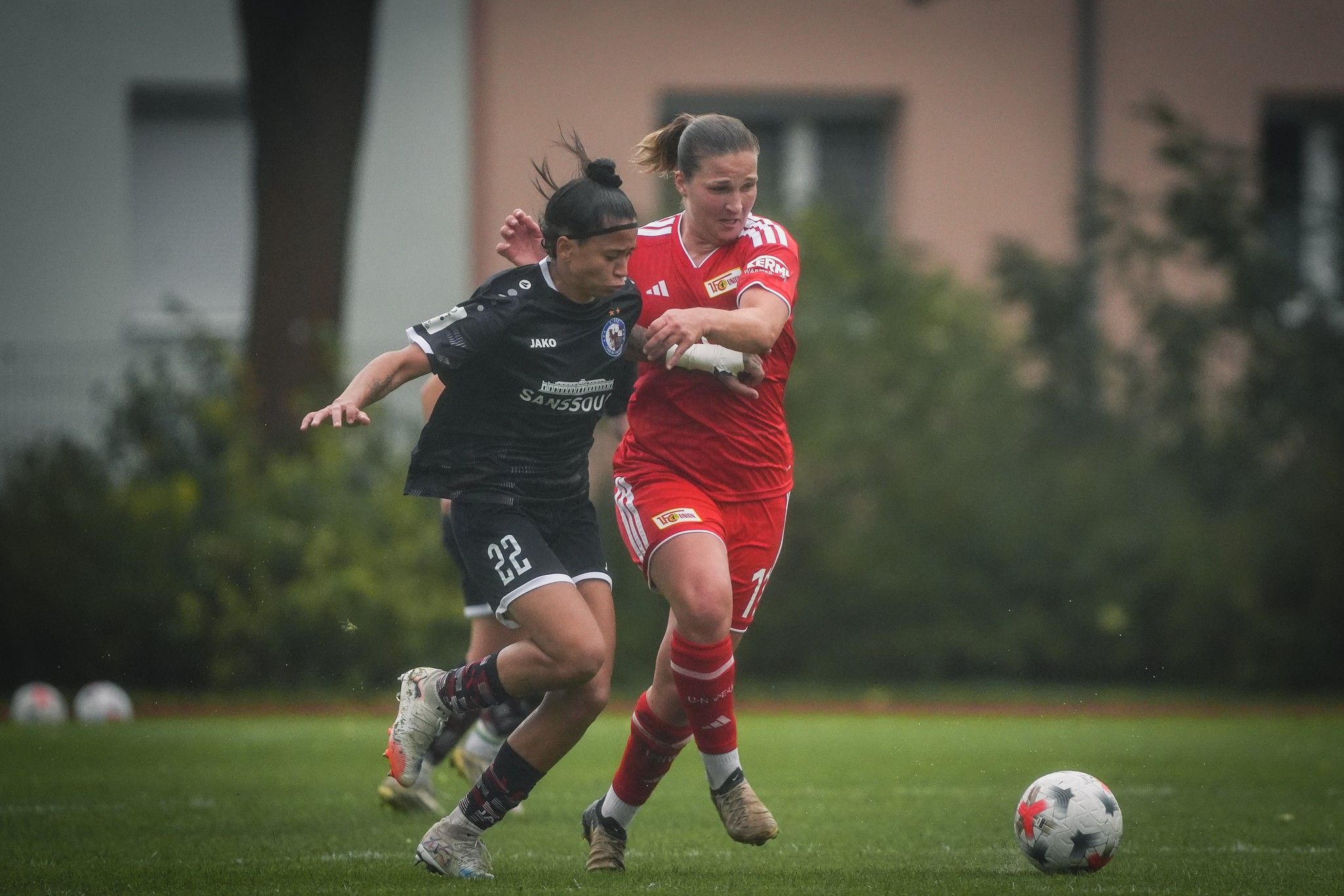Zwei Frauen im Fußballtrikot kämpfen um den Ball auf einem grünen Rasenfeld, im Hintergrund sind Bäume und ein Gebäude sichtbar.