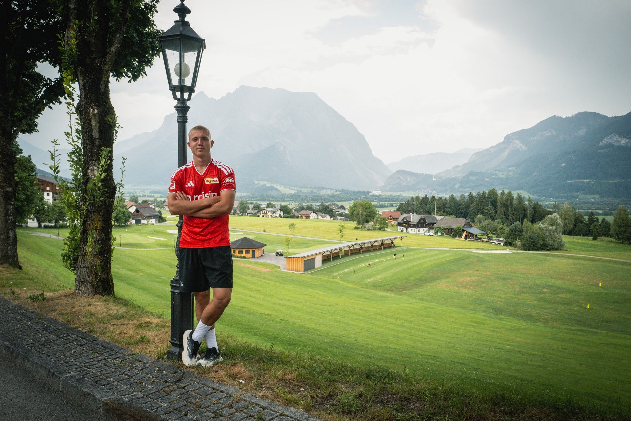 Ein Athlet in rotem Trikot lehnt an einer Laterne, mit Berglandschaft und Sporteinrichtungen im Hintergrund.