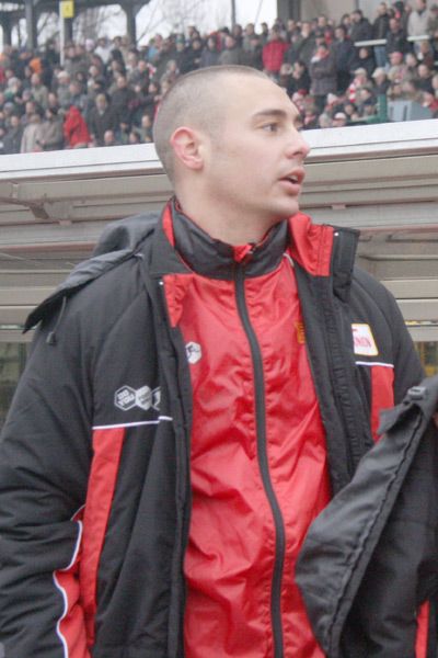 Ein Mann in rot-schwarzer Sportkleidung steht in einer Menschenmenge, vermutlich am Rande eines Sportereignisses.