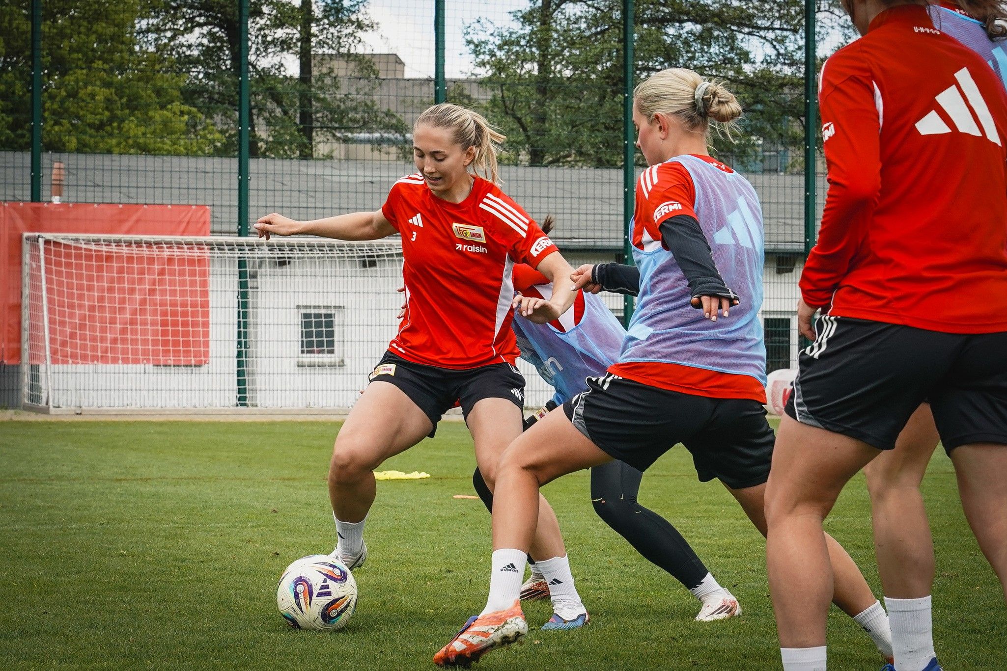 Drei Fußballspielerinnen trainieren auf einem Rasenplatz, während eine Spielerin den Ball dribbelt.