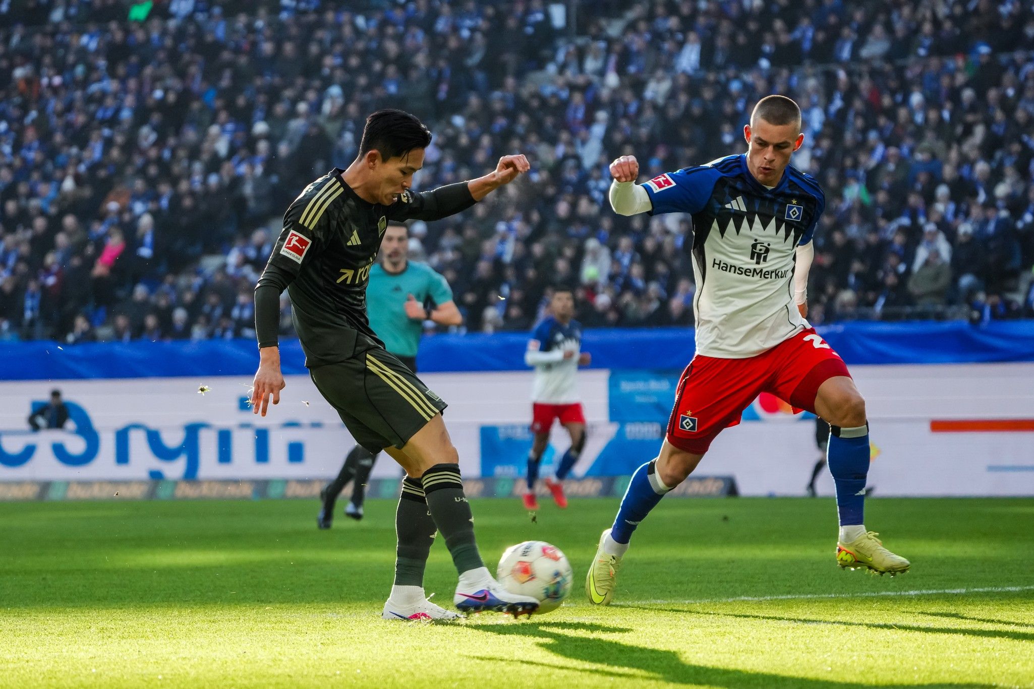 Spielszene im Fußball, ein Spieler dribbelt den Ball, während ein anderer Verteidiger ihn anzugreifen versucht. Zuschauer im Hintergrund.