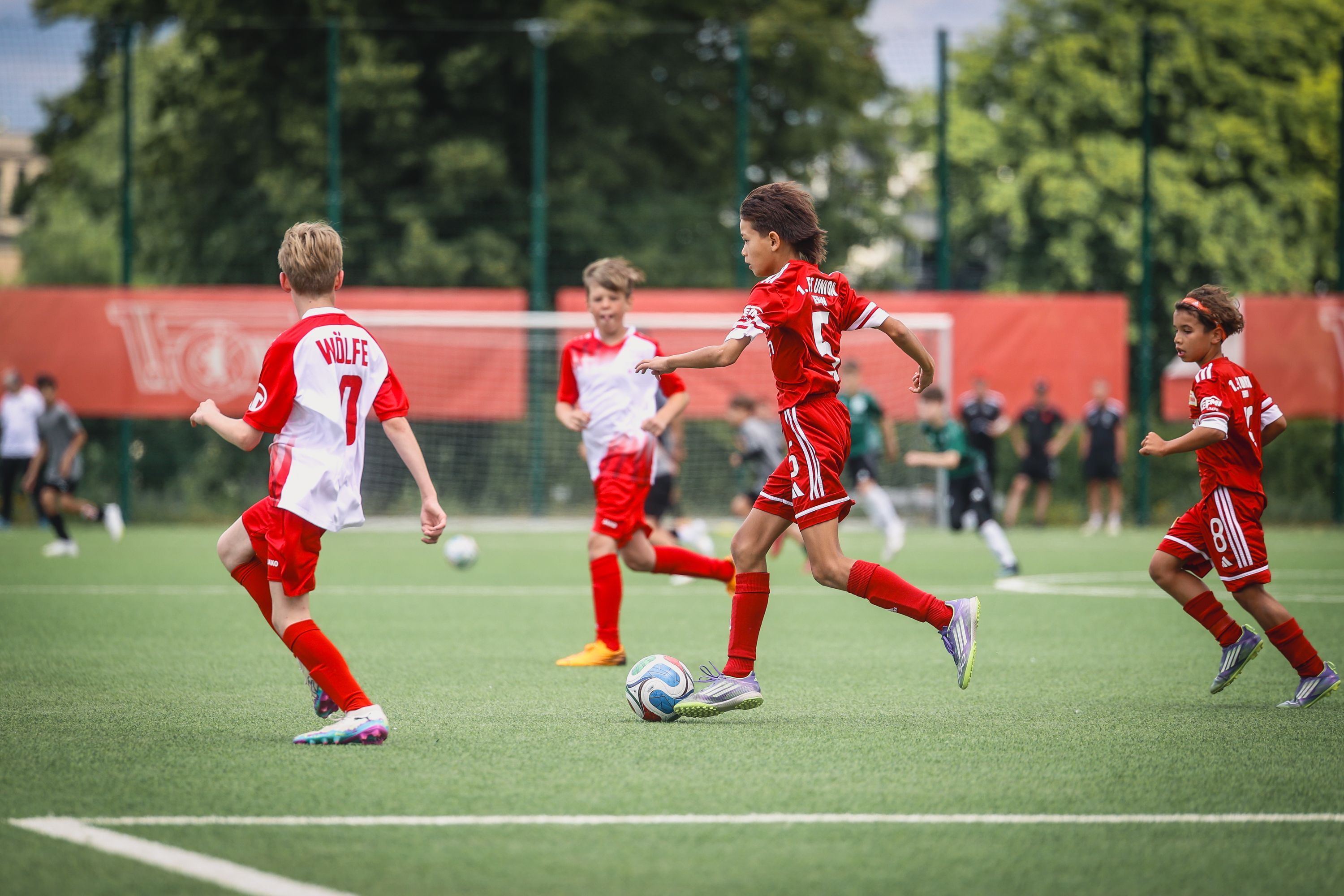 Vier Kinder in Fußballtrikots spielen auf einem Kunstrasenplatz. Zwei Spieler dribbeln den Ball, während andere im Hintergrund stehen.