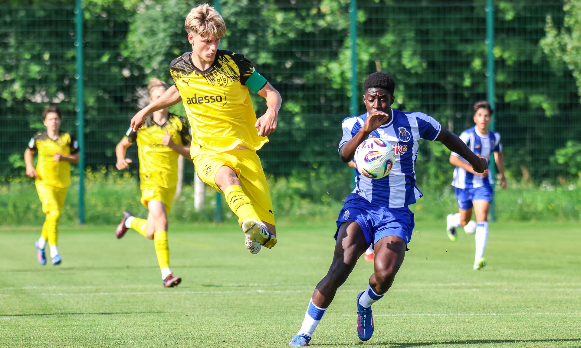 Junger Fußballspieler in gelber Trikotsprung, während ein Spieler in blauem Trikot den Ball führt, auf einem grünen Rasenplatz.