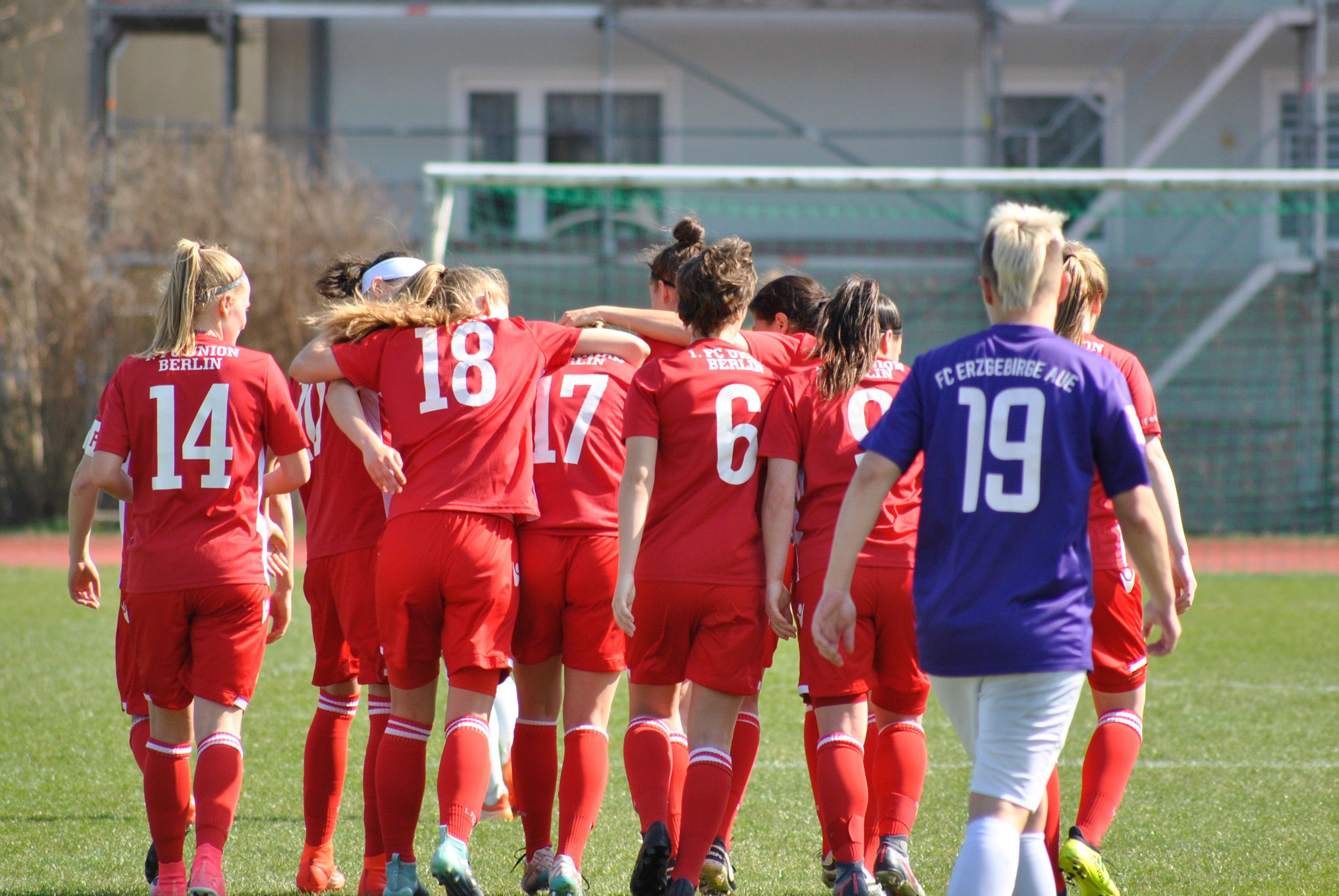 Eine Gruppe Frauen trägt rote Fußballtrikots. Sie umarmen sich auf dem Spielfeld. Im Hintergrund ist ein Tor zu sehen.