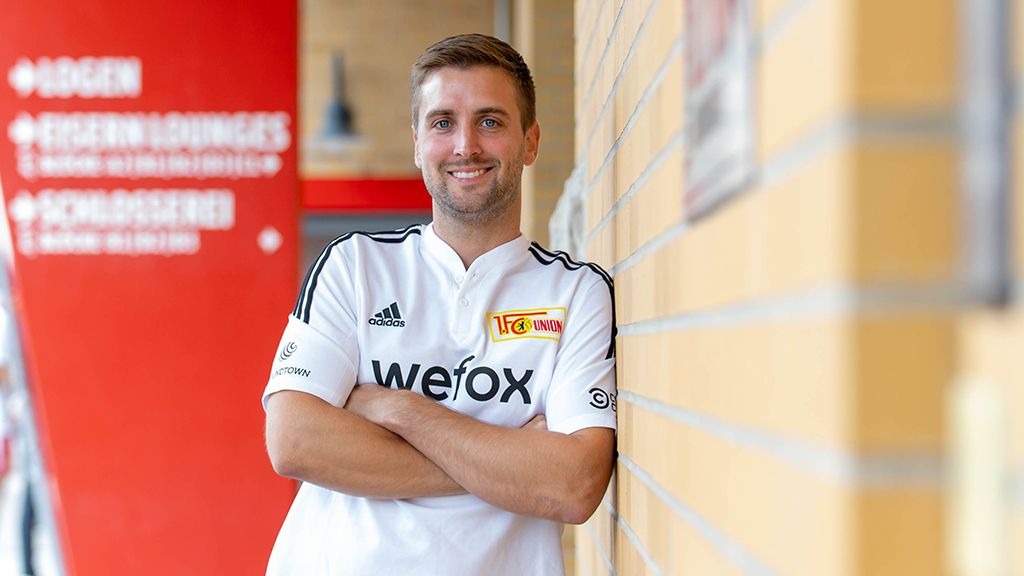 Lächelnder Mann in einem weißen Sporttrikot lehnt an einer Wand, im Hintergrund ein rotes Plakat.