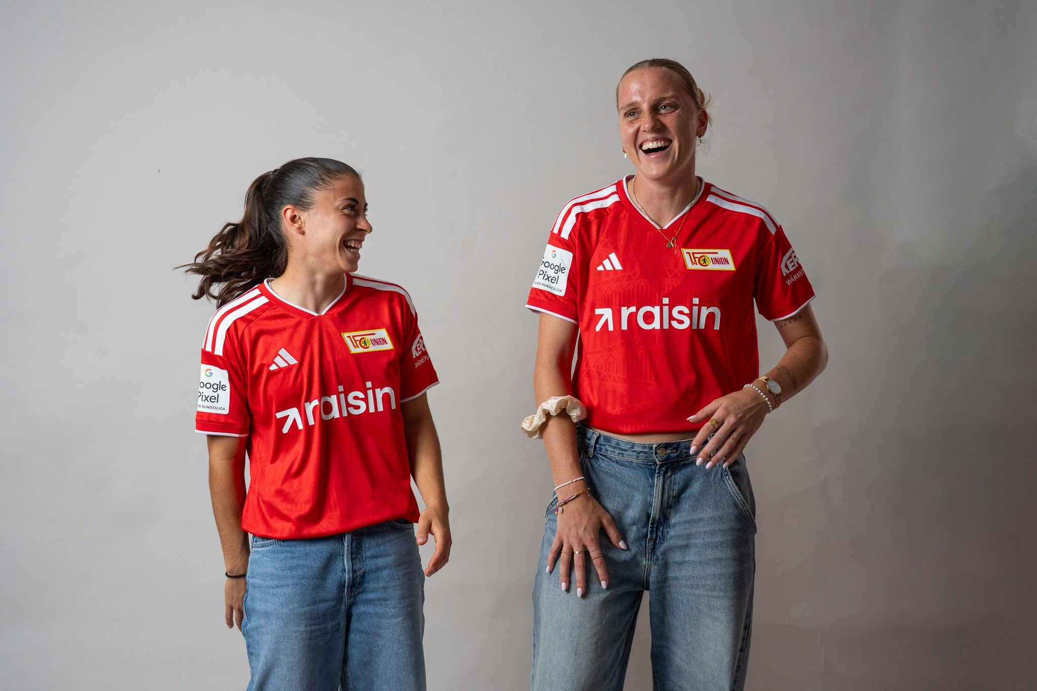 Zwei Lächelnde Frauen in roten Fußballtrikots stehen nebeneinander und lachen, während sie an einem neutralen Hintergrund posieren.