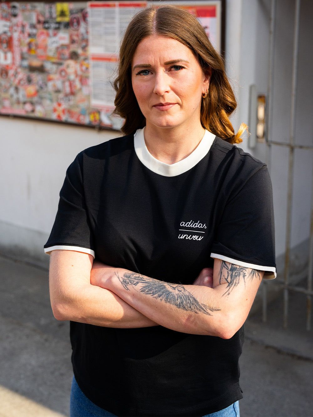 Eine Person mit langen, braunen Haaren steht mit verschränkten Armen in einem schwarzen T-Shirt vor einer Wand mit Plakaten.