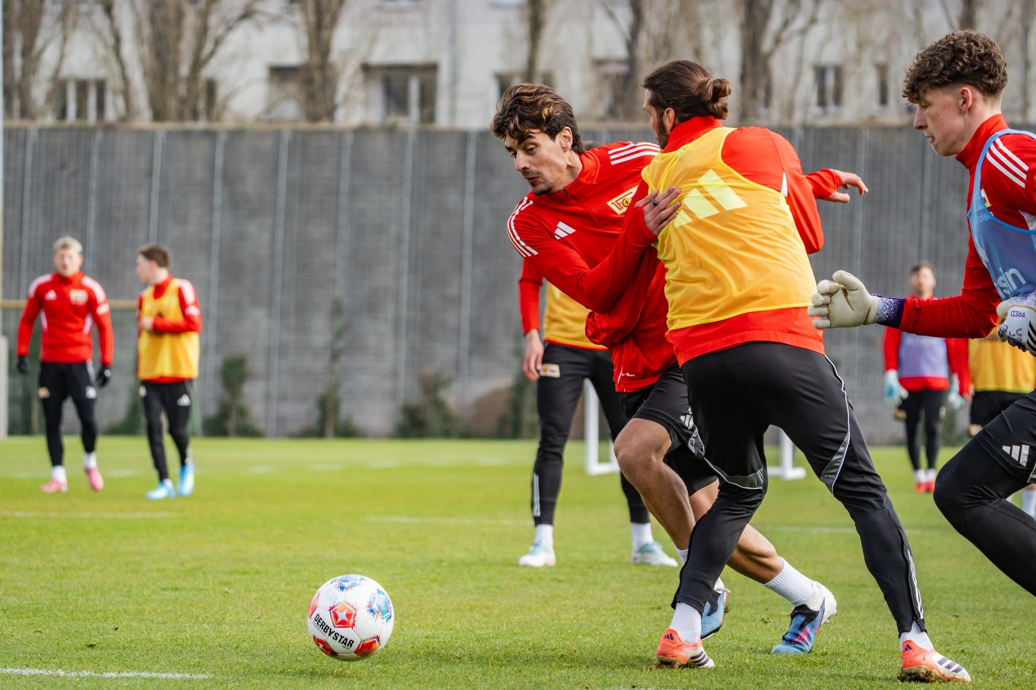 Zwei Fußballspieler im Training: einer mit rotem Trikot dribbelt, während ein anderer in gelbem Shirt ihn verfolgt.