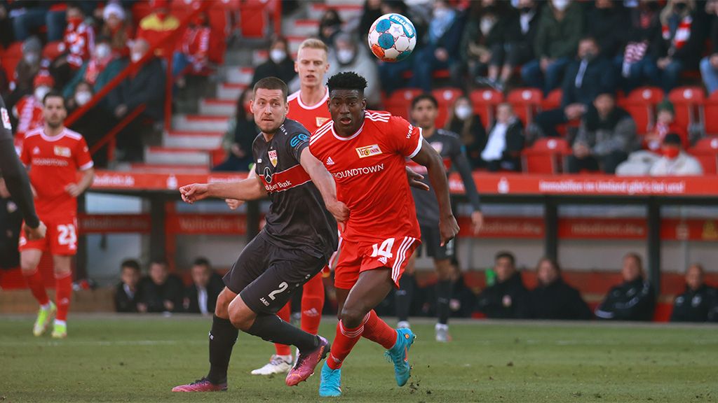 Fußballspiel zwischen zwei Teams, ein Spieler in rotem Trikot mit der Nummer 14 führt den Ball, während ein gegnerischer Spieler in Schwarz drängt.