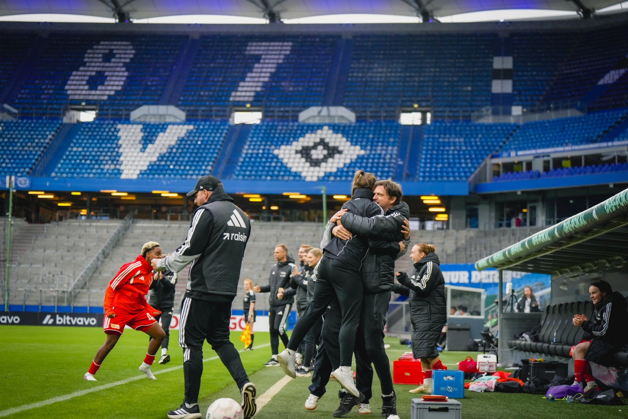 Trainer und Spieler umarmen sich freudig während eines Fußballspiels, Zuschauerbereich im Hintergrund.