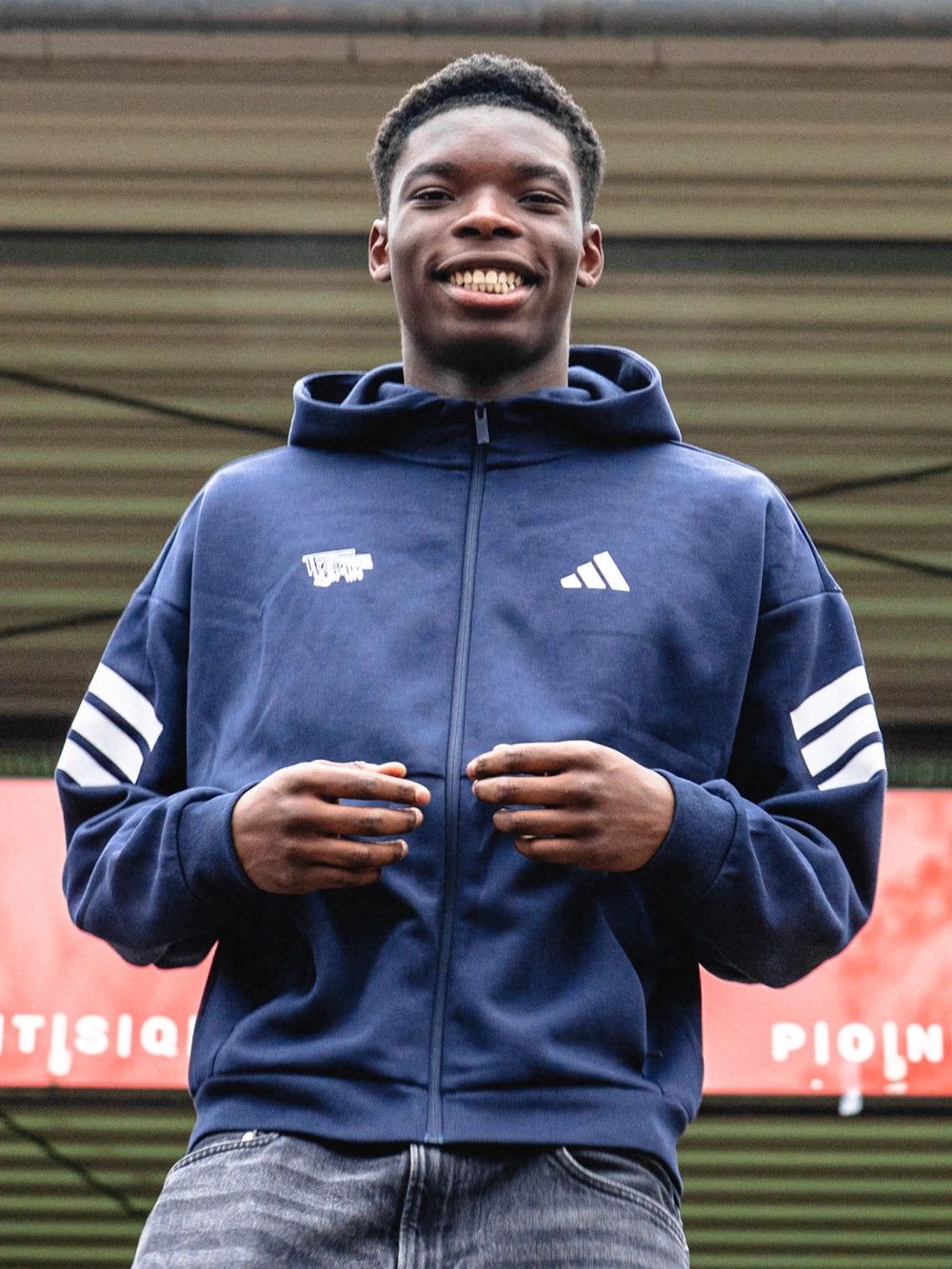 Ein junger Mann in einem blauen Adidas-Hoodie steht lächelnd und posiert vor einer unscharfen Hintergrundkulisse.