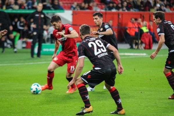 A soccer player dribbles the ball while two defenders try to stop him. Stadium atmosphere in the background.