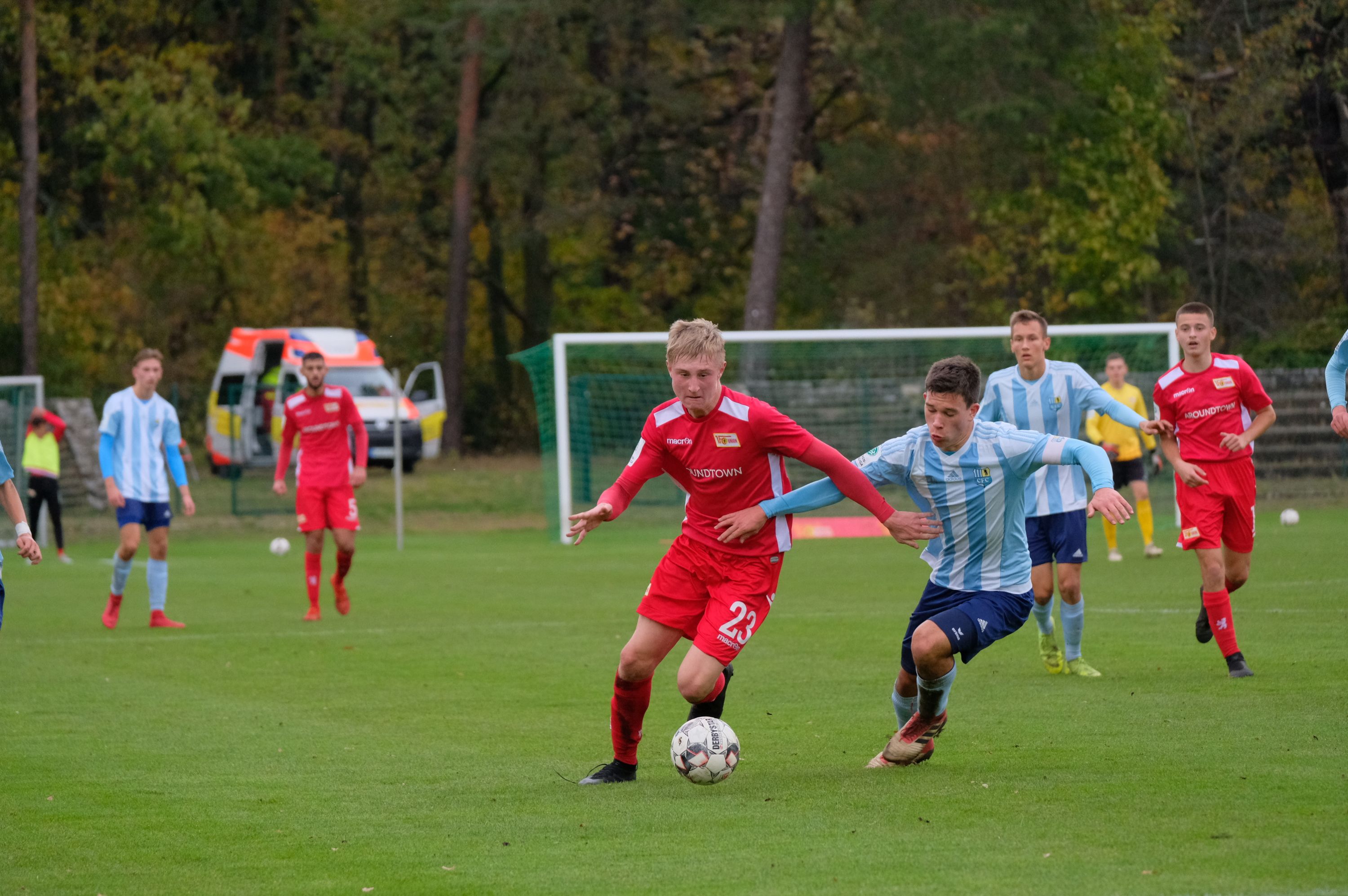 Zwei Fußballspieler kämpfen um den Ball. Sie sind auf einem grünen Platz. Im Hintergrund stehen andere Spieler.