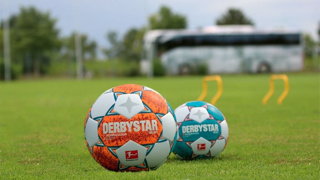 Zwei Fußbälle der Marke Derbystar auf einem grünen Fußballfeld, im Hintergrund ein Bus und einige Trainingsmarkierungen.