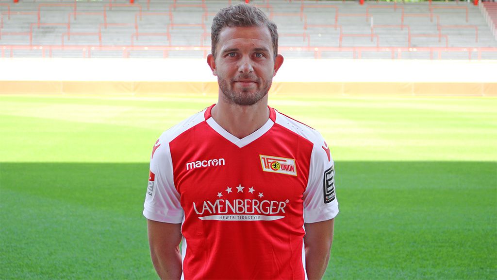 A player stands on a football field wearing a red jersey with a club logo.