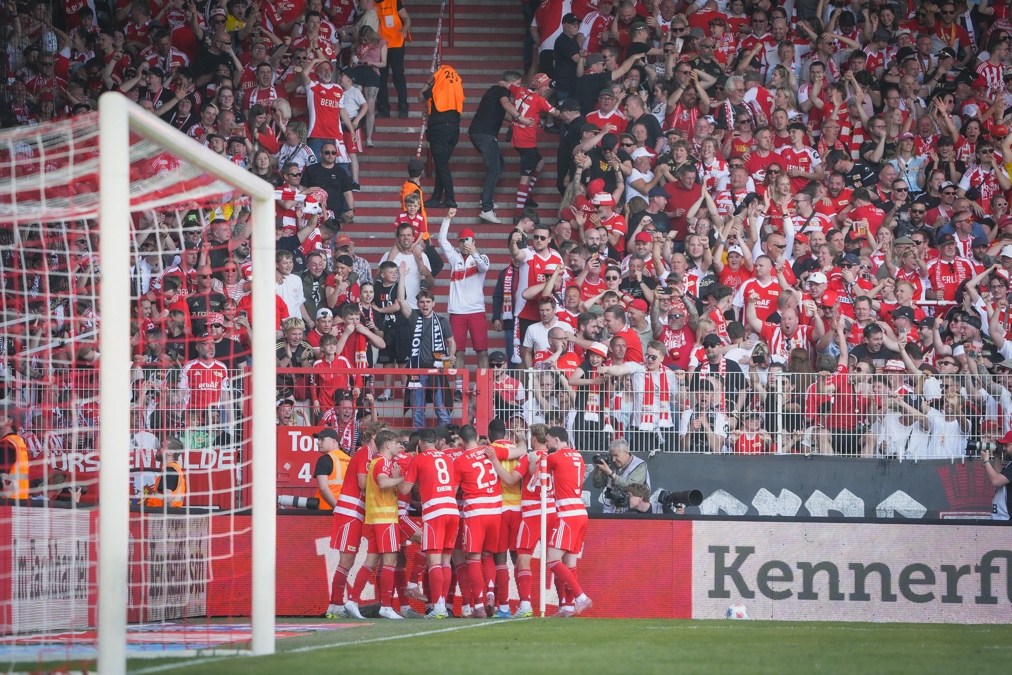 Spieler einer Fußballmannschaft feiern ein Tor vor jubelnden Fans in einem Stadion, das in rot und weiß dekoriert ist.