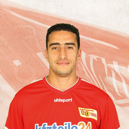 Ein junger Mann mit dunklen Haaren trägt ein rotes Fußballtrikot mit dem Logo eines Vereins im Hintergrund.