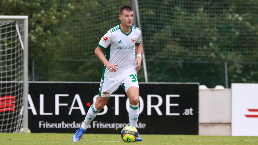 Ein Fußballspieler in einem weißen Trikot mit grünen Akzenten stellt sich auf dem Spielfeld auf. Hinweise auf Werbung im Hintergrund.