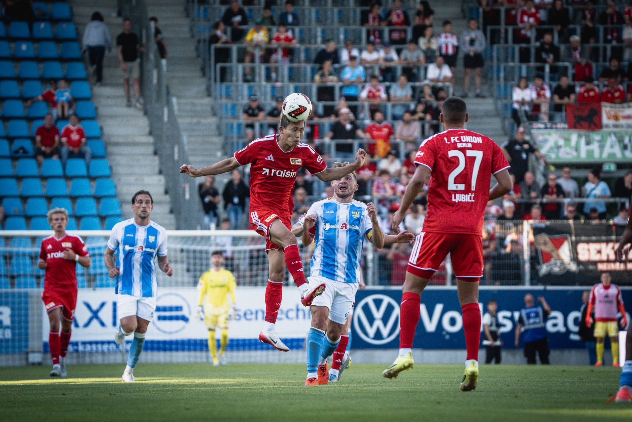 Spielszene im Fußball, ein Spieler springt, um den Ball mit dem Kopf zu spielen, während zwei Gegenspieler in der Nähe stehen.