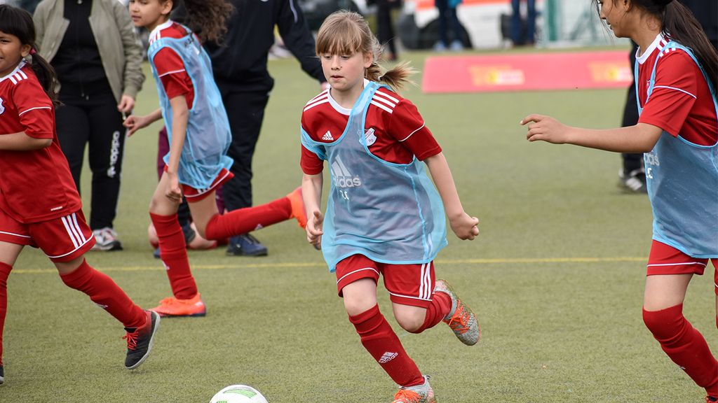 Mädchen in roter Sportkleidung und blauer Weste spielt Fußball auf einem Kunstrasenfeld, umgeben von anderen Kindern.