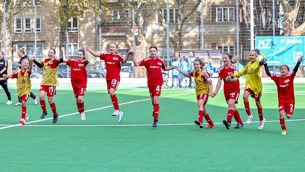 Mädchenfußballmannschaft in roten Trikots feiert jubelnd auf dem Spielfeld, einige Spielerinnen halten sich an den Händen.