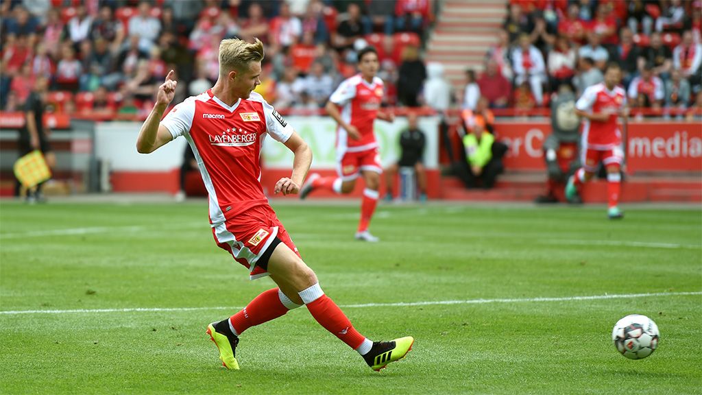 Spielszene eines Fußballspiels, in der ein Spieler in roter Kleidung einen Schuss aufs Tor abgibt, im Hintergrund weitere Spieler.