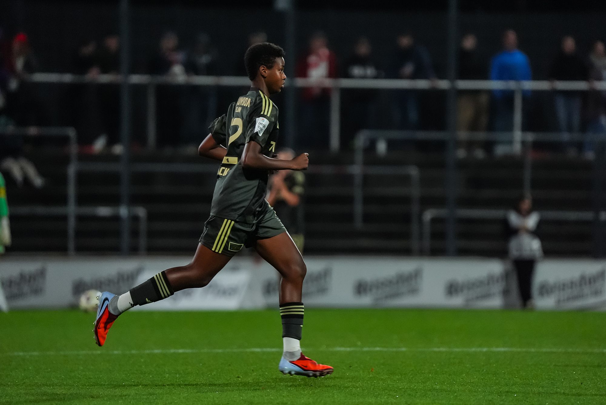 Ein junger Fußballspieler in einem dunkelgrauen Trikot läuft auf einem Fußballplatz während eines Spiels. Zuschauer sind im Hintergrund zu sehen.