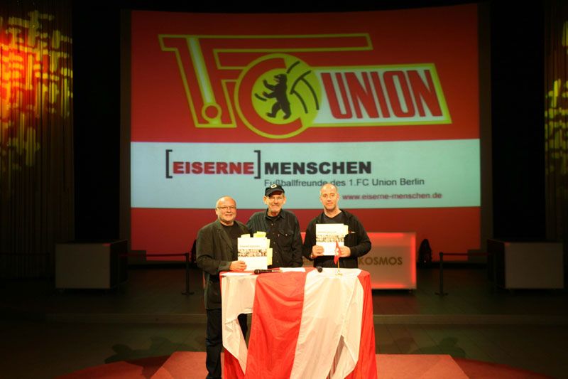 Drei Personen stehen an einem Tisch mit Urkunden vor einer großen Präsentationsleinwand mit dem Logo des 1. FC Union Berlin.