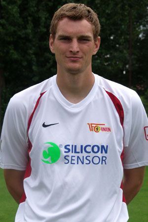 Ein junger Mann in einem weißen Fußballtrikot mit Logos, steht im Freien vor einem grünen Hintergrund.