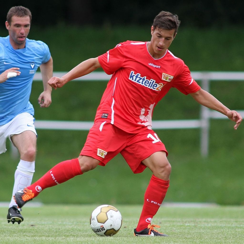 Zwei Fu ßballspieler auf dem Platz: einer in rotem Trikot mit Ball, der andere in blauem Trikot, im Hintergrund.