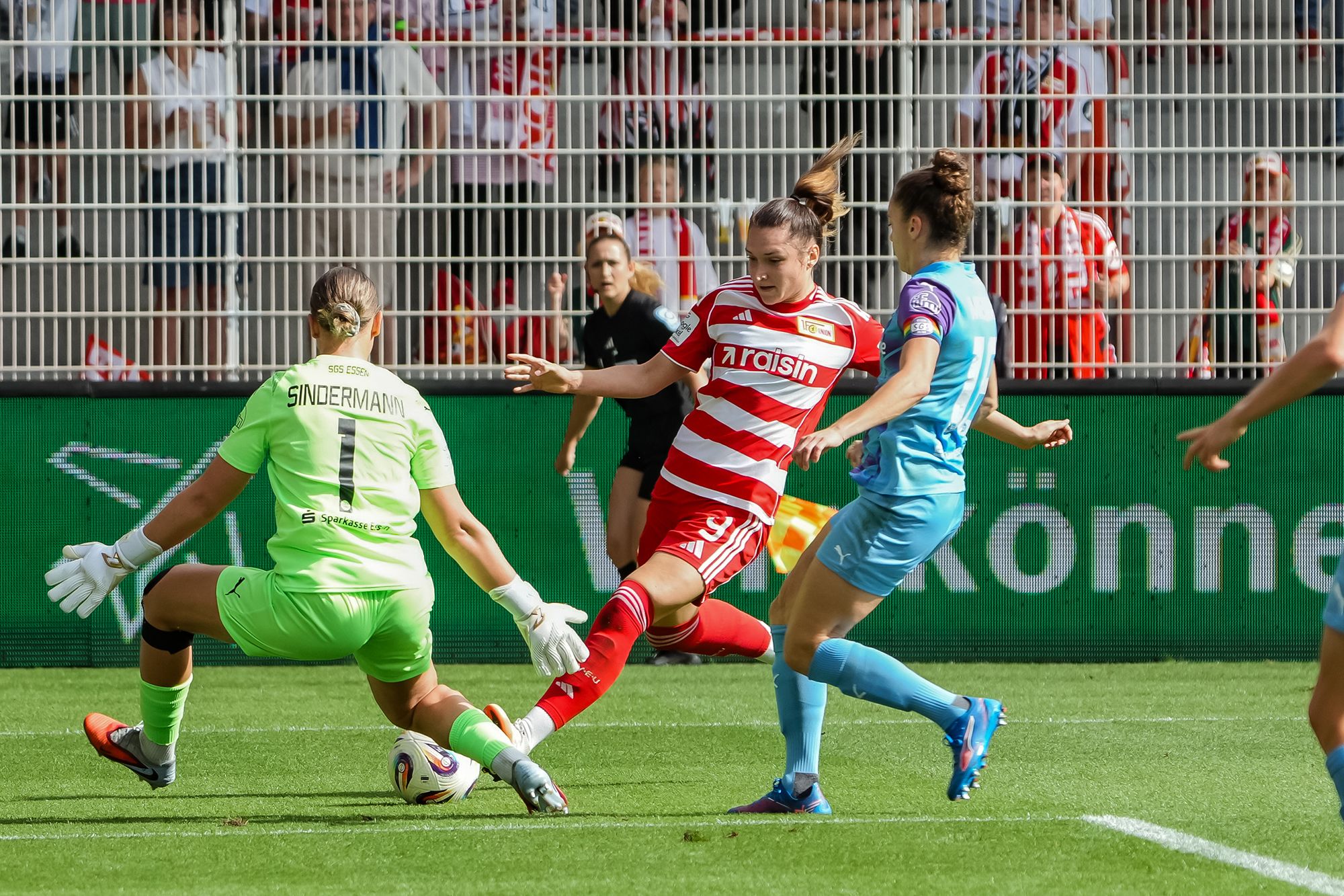 Eine Spielerin in rotem Trikot dribbelt den Ball, während eine Torhüterin in Grün und eine Spielerin in Blau sie angreifen.