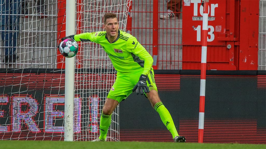 Torwart in gelber Ausrüstung steht im Tor, bereit, einen Ball zu fangen. Im Hintergrund ist das Tor und die Tribüne sichtbar.