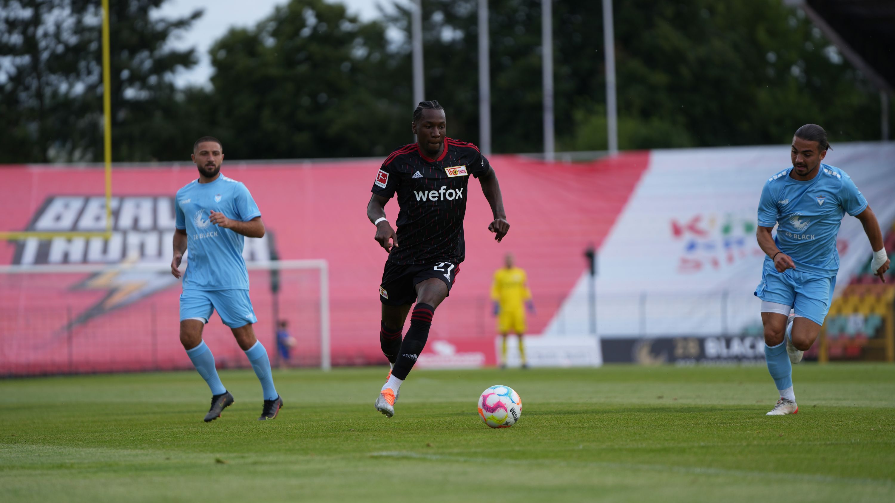 Ein Fußballspieler trägt schwarz und rot. Er dribbelt den Ball. Zwei Spieler in hellblau folgen ihm. Im Hintergrund ist ein Fußballtor.