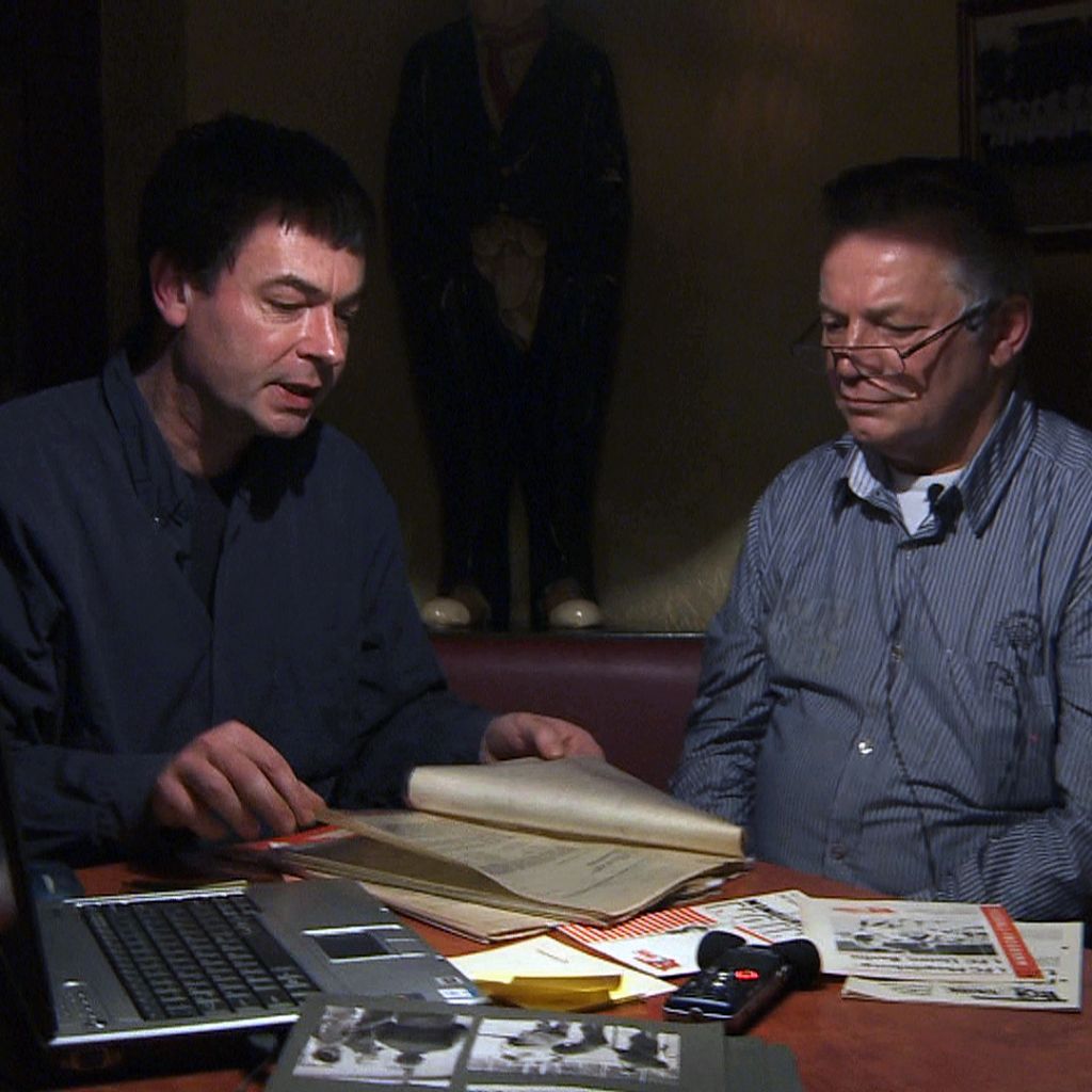 Zwei Männer sitzen an einem Tisch, betrachten Dokumente und Notizen, während ein Laptop und verschiedene Unterlagen sichtbar sind.