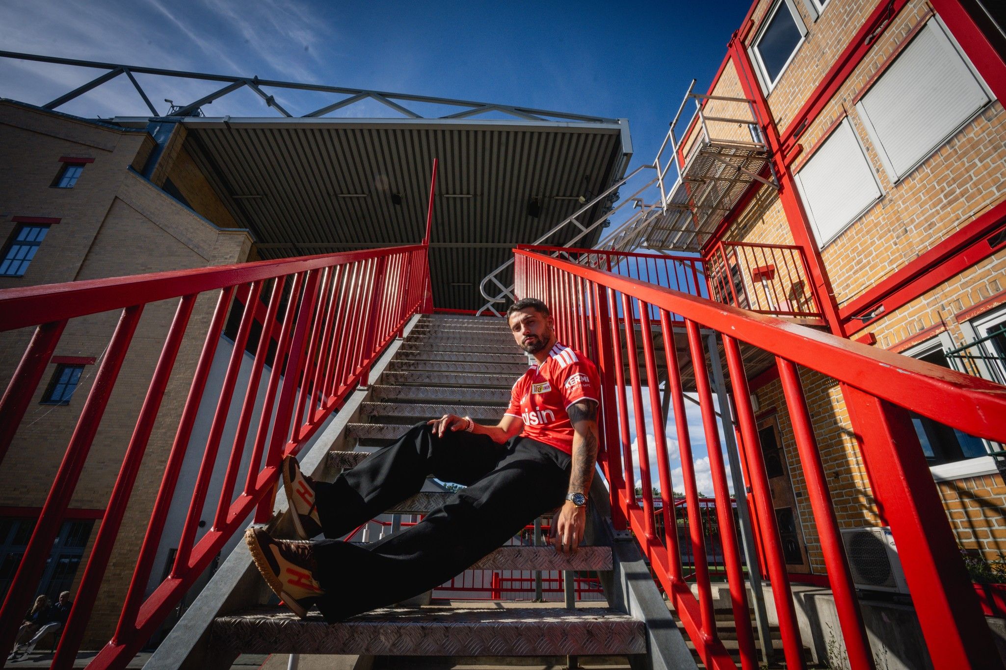 Junger Mann im roten Trikot sitzt auf einer Treppe, umgeben von Gebäuden und unter blauem Himmel.