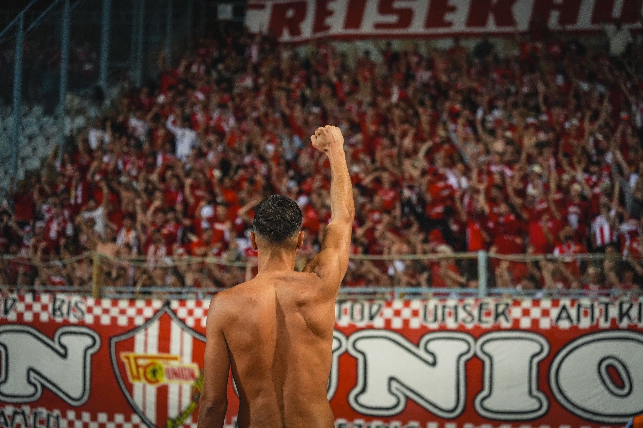 Ein jubelnder, oberkörperfreier Fußballspieler hebt die Faust in die Luft vor einer jubelnden Fangruppe in einem Stadion.