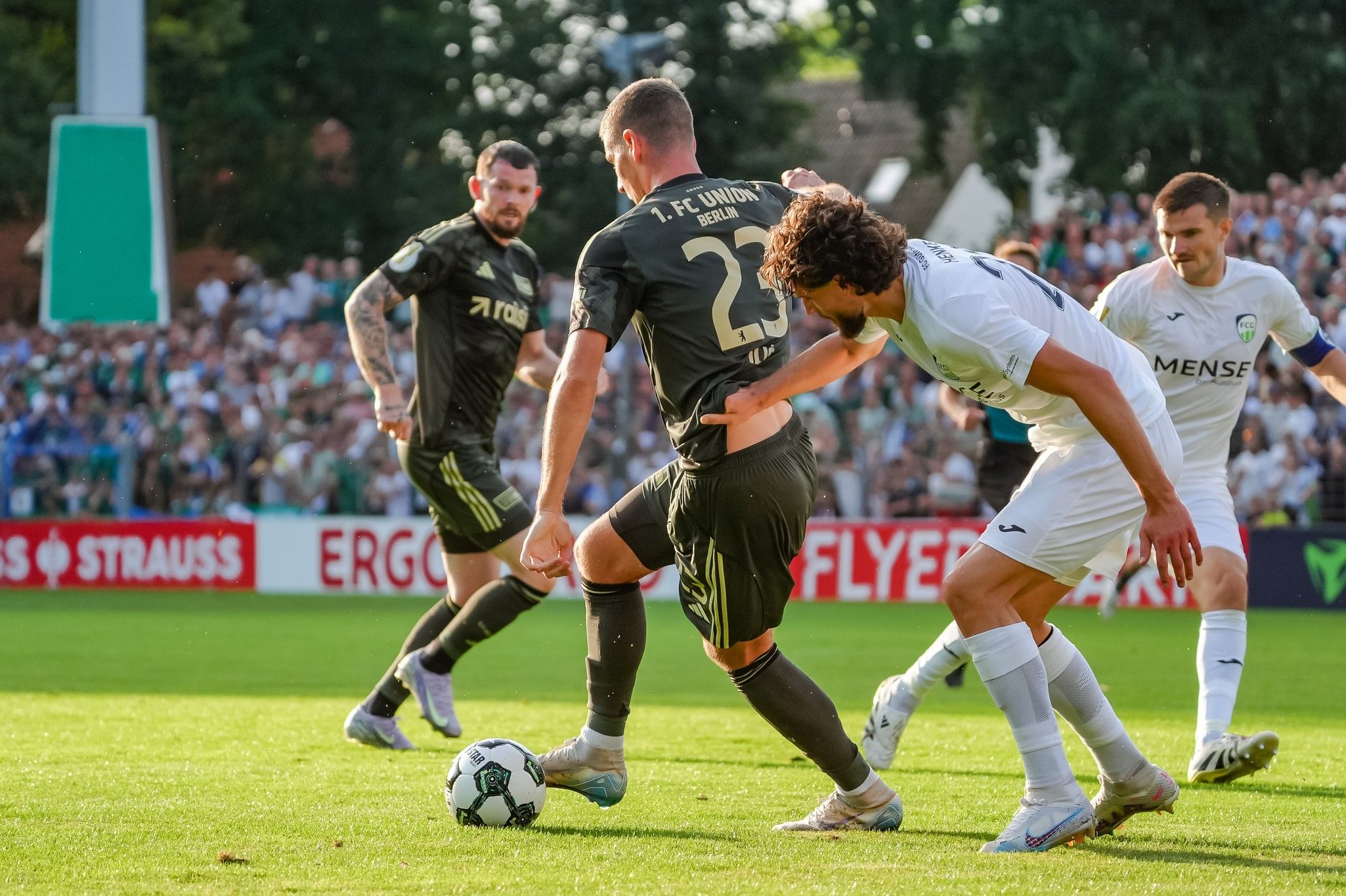 Fußballspiel mit Spielern in schwarzer und weißer Ausrüstung, die um den Ball kämpfen. Zuschauer im Hintergrund.
