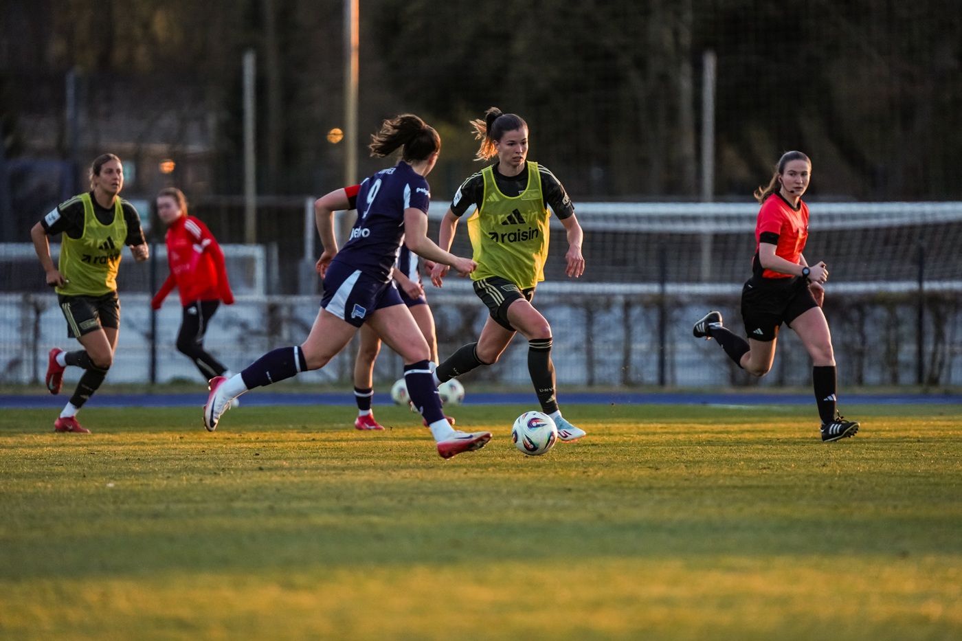 Fünf Spielerinnen im Wettkampf auf einem Fußballfeld, im Hintergrund Tore und Zuschauer.