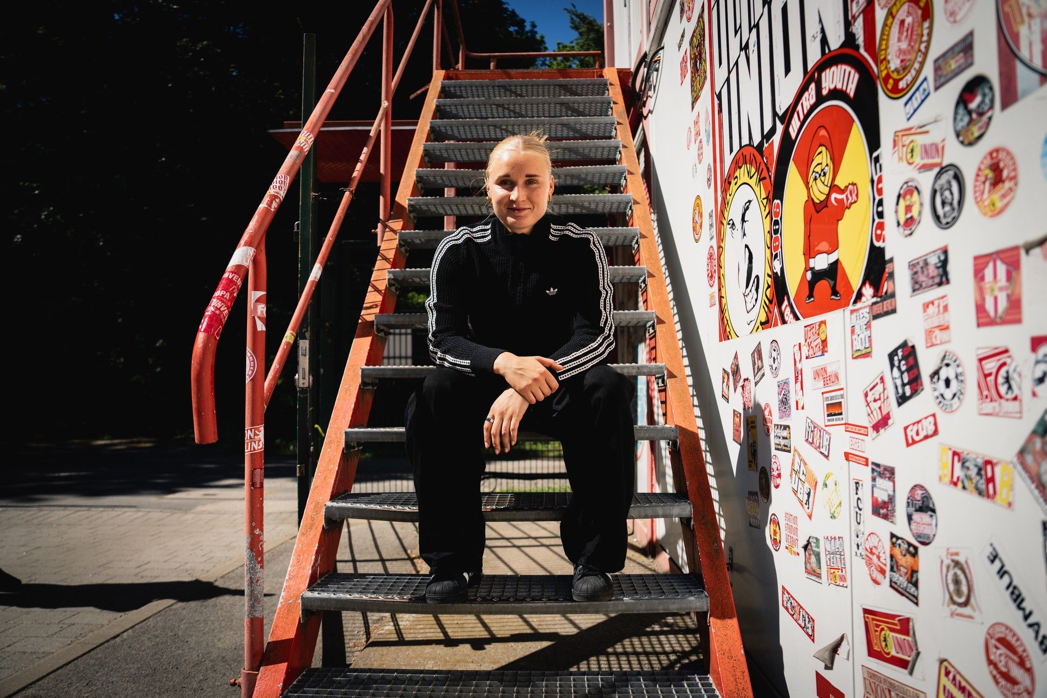 Eine Person sitzt auf einer Treppe, trägt sportliche Kleidung und steht vor einer Wand mit Aufklebern und Grafiken.