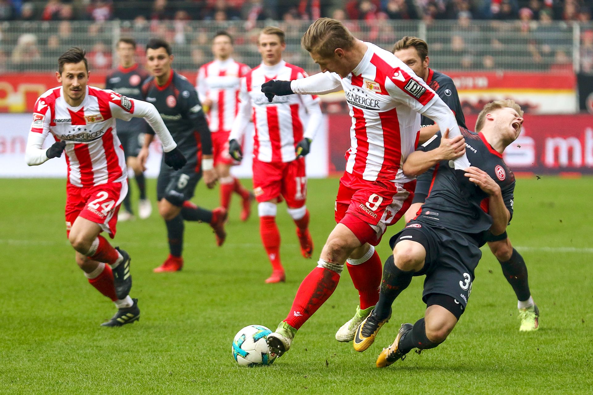 Ein Fußballspieler trägt ein rotes und weißes Trikot. Er kämpft um den Ball. Ein anderer Spieler trägt ein schwarzes Trikot. Er fällt.
