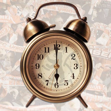 Ein Wecker mit einem goldenen Gehäuse und großen Ziffern, vor einem Hintergrund aus Fanschals und -bildern.