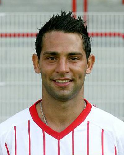 Porträt eines lächelnden Mannes mit kurzen, spiky Haaren, gekleidet in ein weißes Fußballtrikot mit roten Streifen.