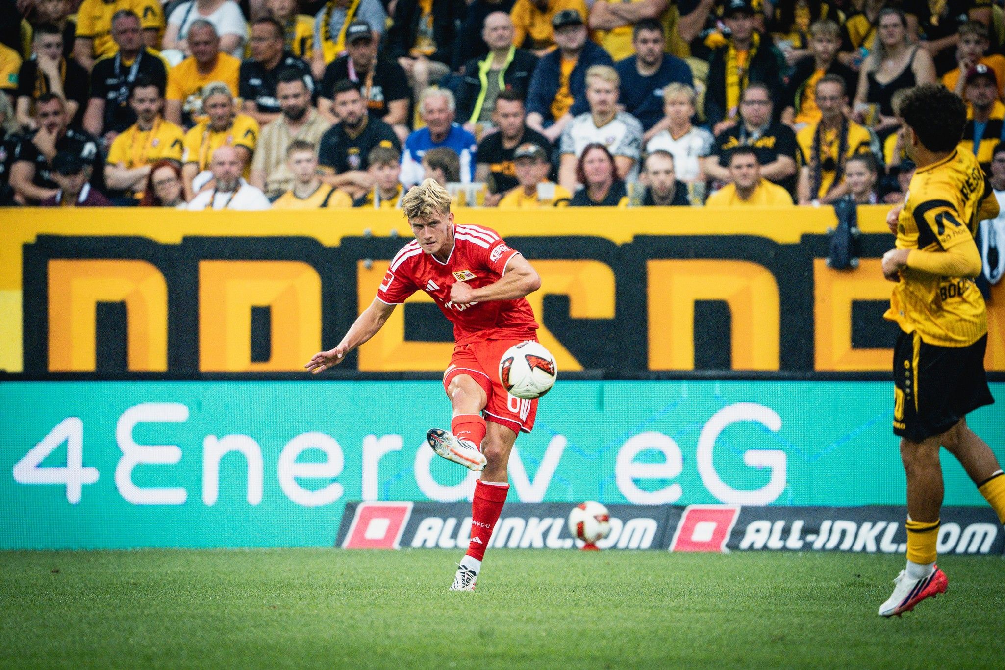 Fußballspieler in rotem Trikot tritt den Ball im Stadion, im Hintergrund jubelnde Fans in gelben Trikots.