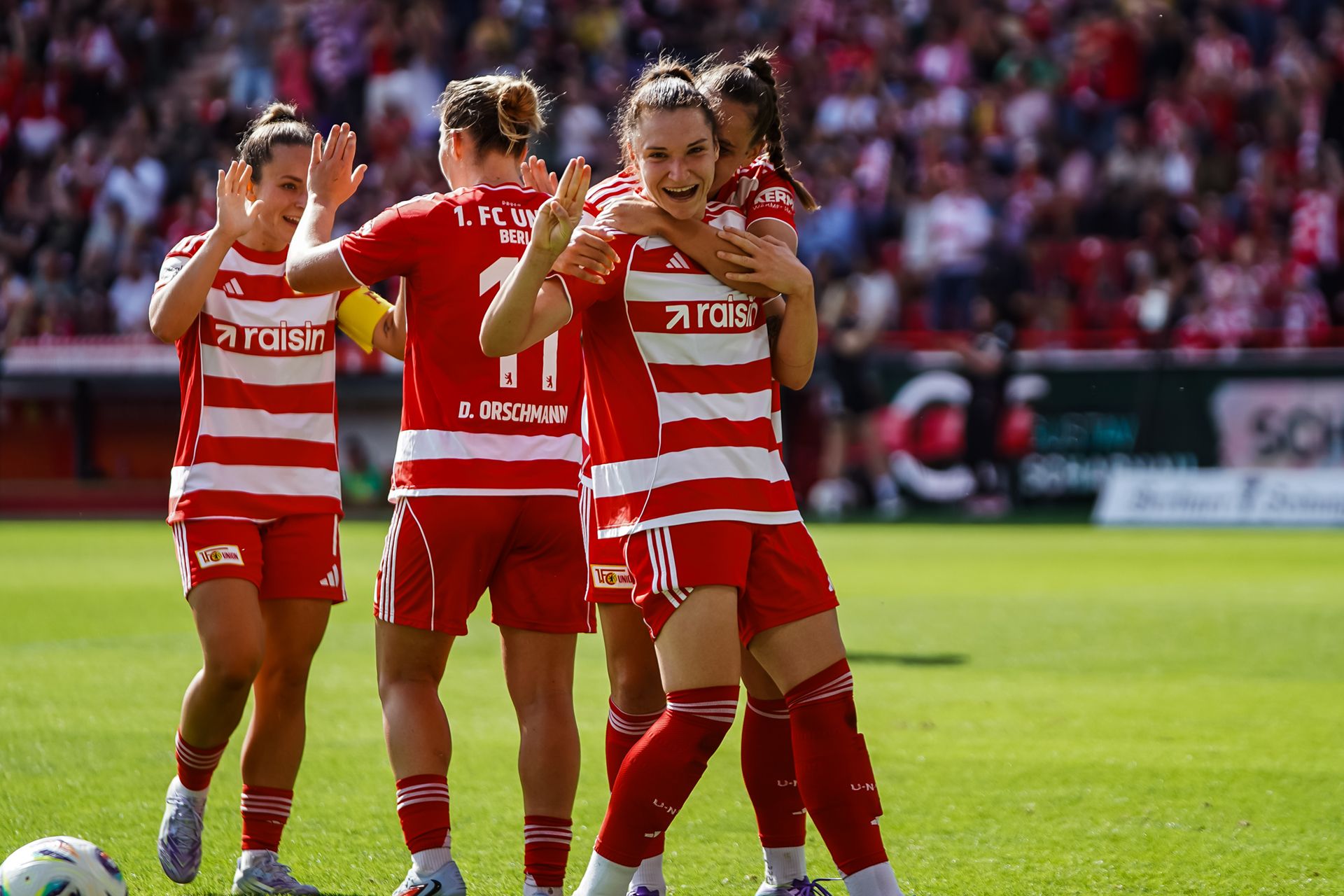 Fußballspielerinnen in roten Trikots feiern ein Tor, umgeben von jubelnden Fans im Stadion.